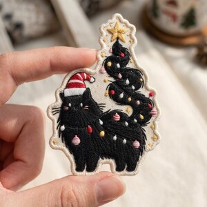 May include: An embroidered patch featuring a fluffy black cat wearing a red and white striped Santa hat. The cat's long, winding tail forms the shape of a Christmas tree, decorated with small colorful ornaments and topped with a gold star.