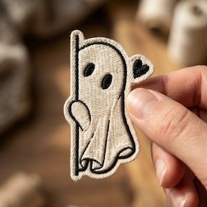 May include: An embroidered patch featuring a cream-colored ghost peeking from behind a vertical black line. The ghost has two oval black eyes and a small black heart shape on its right side, all outlined in thick black thread stitching.
