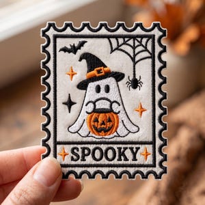 May include: A rectangular embroidered patch shaped like a postage stamp with a white background and black scalloped border. It features a white ghost wearing a black witch hat, holding an orange jack-o'-lantern, with a bat, spider, and the word SPOOKY.