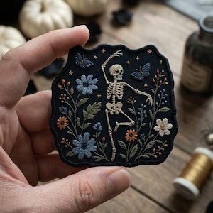 May include: A dark navy blue embroidered patch featuring a central off-white skeleton in a dancing pose, surrounded by light blue and orange flowers, green leaves, and two blue butterflies, all accented with small gold-colored embroidered stars.