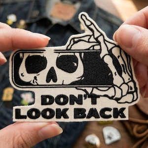May include: An embroidered patch featuring a black and white design of a skeleton hand holding a rearview mirror with a skull reflection. The text DON'T LOOK BACK is stitched in black thread on a cream-colored background with a die-cut border.