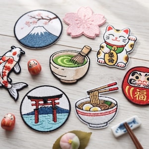 May include: A collection of embroidered patches featuring Japanese motifs including Mount Fuji, a cherry blossom, a beckoning cat, a koi fish, a matcha bowl with whisk, a torii gate, a ramen bowl with chopsticks, and a red Daruma doll.