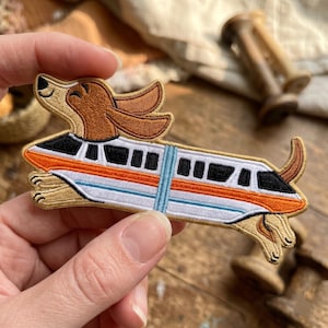 May include: An embroidered patch featuring a brown and tan dog with long ears integrated into the body of a white monorail train. The train has black windows, an orange stripe, and a light blue vertical accent line, with the dog's head and tail extending from the ends of the train car.