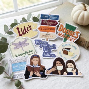 May include: A collection of embroidered patches featuring various designs including a coffee cup labeled Luke's, a directional signpost, a Doose's Market logo, a dragonfly, the text Where you lead, I will follow, and illustrations of people with books and coffee.
