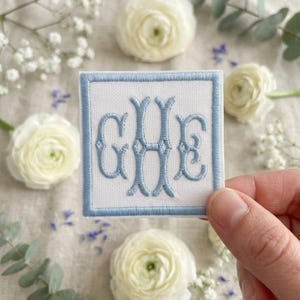 May include: A square white fabric patch featuring a light blue embroidered border and a central light blue monogram with the letters G, H, and E in a decorative script font. The patch is held against a background of white flowers and greenery.