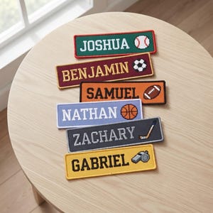 May include: Six rectangular embroidered name patches featuring sports themes. The patches display the names JOSHUA, BENJAMIN, SAMUEL, NATHAN, ZACHARY, and GABRIEL in various colors including green, maroon, orange, light blue, dark gray, and yellow.