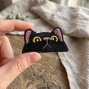 May include: An embroidered patch shaped like a black cat head with a flat bottom edge. The cat features bright yellow eyes, a light gray nose, and pink inner ears, all rendered in dense black thread with a distinct stitched border outline.