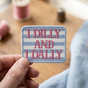 May include: A rectangular embroidered patch featuring horizontal light blue and white stripes. The text I DILLY AND I DALLY is stitched in a bold pink thread across the center of the patch, which has a finished stitched border in light blue.