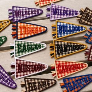 May include: A collection of triangular pennant-shaped embroidered patches featuring various team names like Wildcats, Tigers, Bulldogs, Eagles, Panthers, Mustangs, Cardinals, Bears, and Gators, each with a distinct gingham check pattern and fringe.