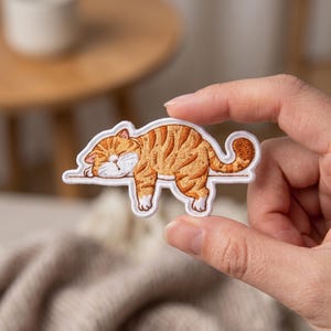 May include: An embroidered patch featuring a sleeping orange tabby cat with dark stripes and a white face, chest, and paws. The cat is depicted lying horizontally on a thin white line, with its tail curled upward and its eyes closed in rest.