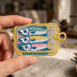 May include: An embroidered patch shaped like an open rectangular tin can containing three stylized fish. The fish are colored in shades of green, yellow, and pink with blue details. The tin has a yellow border and a silver pull-tab mechanism.