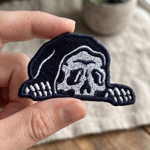 May include: An embroidered patch featuring a dark navy blue hooded figure with a white skull face and skeletal hands peeking out from the bottom edge. The design is rendered in high-contrast thread with detailed stitching on the skull and hood.