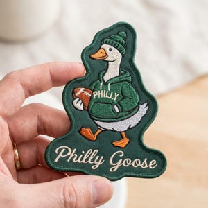 May include: An embroidered patch featuring a white goose wearing a green beanie and a green hoodie with the word PHILLY printed on it. The goose holds a brown football. Below the goose, the text Philly Goose is embroidered in white script.