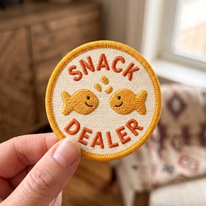 May include: A circular embroidered patch with an off-white background and a golden-yellow border. The patch features two orange fish facing each other with small orange bubbles above them and the text SNACK DEALER in orange embroidery.