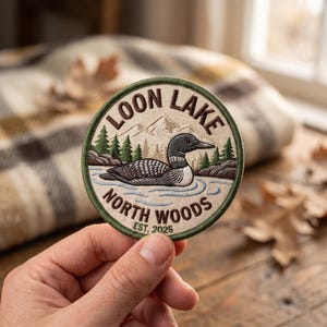 May include: A circular embroidered patch featuring a loon swimming on water with green pine trees and mountains in the background. The text reads LOON LAKE NORTH WOODS EST. 2025 in dark brown lettering. The patch has a dark green stitched border.