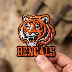 May include: An embroidered patch featuring the head of a tiger with orange, black, and white thread. Below the tiger head, the word BENGALS is stitched in orange capital letters with a black border, all set against a black fabric background.