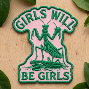 May include: An embroidered patch with a light pink background and a dark green border. The patch features a green praying mantis illustration and the dark green embroidered text GIRLS WILL BE GIRLS, with the word GIRLS at the top and BE GIRLS below.