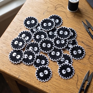 May include: A collection of circular black embroidered patches featuring a white scalloped border and two large white circular eyes with black pupils. The patches are scattered across a wooden surface alongside black thread and metal sewing scissors.