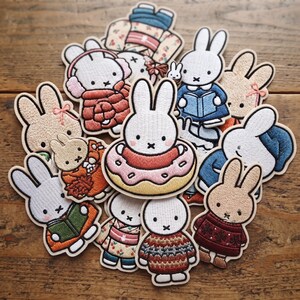 May include: A collection of embroidered fabric patches featuring Miffy the rabbit in various outfits and poses, including wearing a sweater, reading a book, and sitting inside a pink donut. The patches are white, tan, and multi-colored.
