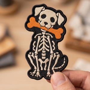 May include: An embroidered patch featuring a dog skeleton in cream thread on a black background. The dog is sitting and holding a bright orange bone in its mouth. The entire design is outlined with a thick black border, creating a distinct shape.