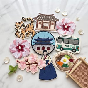 May include: A collection of embroidered patches featuring a tiger, a traditional building, two pink hibiscus flowers, a circular palace scene, a green Seoul BUS, a cherry blossom branch, a person in a hanbok, and a bowl of bibimbap with chopsticks.