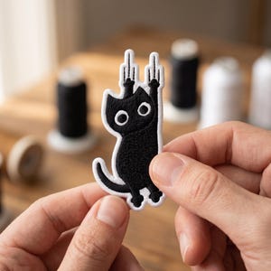 May include: An embroidered patch featuring a black cat with large white eyes, shown with both front paws raised and extended upward, each displaying a middle finger gesture. The cat has a white border outline and a curved tail at the bottom.