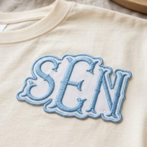 May include: A cream-colored t-shirt featuring a large, central embroidered patch. The patch displays the letters SEN in a light blue, serif-style font, outlined with a matching blue border against a white, textured fabric background.