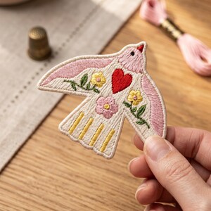 May include: An embroidered bird patch featuring a cream-colored body with pink wings, a red heart, and yellow and pink floral accents. The bird has green stems and leaves, with yellow vertical stripes on its tail feathers, all outlined in cream thread.