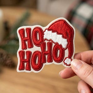 May include: An embroidered patch featuring the red text HO HO HO with a red and white Santa hat integrated into the design. The patch is set against a light cream-colored fabric background with a visible woven texture and a die-cut border.