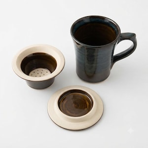 May include: A ceramic tea mug with a dark blue and brown glazed finish, accompanied by a matching tea infuser basket with a perforated base and a circular lid, both featuring a cream-colored rim and a dark brown interior glaze.