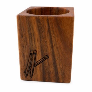 May include: A square wooden pencil holder with a natural brown wood grain finish. The front face features a dark, laser-engraved graphic of three pencils arranged in a cluster. The container has a circular opening at the top for storage.