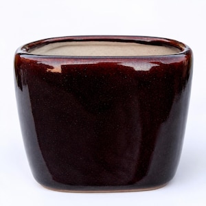 Puede incluir: Una maceta de cerámica de forma cuadrada con esquinas redondeadas y un esmalte brillante de color marrón oscuro profundo. El interior del recipiente es de un color beige mate claro, que contrasta con el acabado exterior oscuro del contenedor.