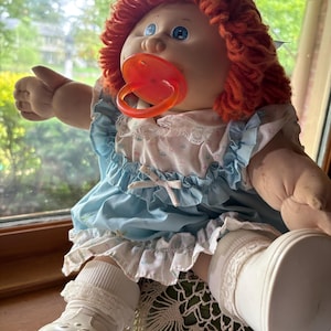 Op de afbeelding: Een pop met fel oranje wollen haar en blauwe ogen, gekleed in een lichtblauwe jurk met een witte rucheskraag en kanten afwerking. De pop heeft een fel oranje fopspeen in de mond en draagt witte sokken en witte plastic schoenen.