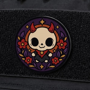 May include: A circular patch featuring a cartoon character with a pale skull head, red horns, and a black outfit. The character is centered against a dark purple background with red flowers, yellow star accents, and a circular orange border.