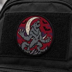 May include: A circular patch featuring a grey werewolf howling at a crescent moon against a dark red background with black silhouettes of bats. The patch is attached to a black fabric surface with a hook-and-loop fastener backing.