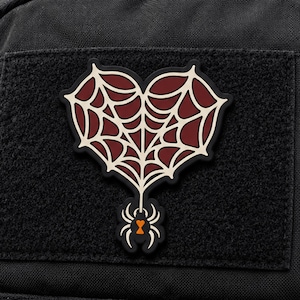May include: A black, heart-shaped patch featuring a cream-coloured spiderweb design against a dark red background. A small black spider with a bright orange hourglass marking on its abdomen hangs from the bottom point of the webbed heart shape.