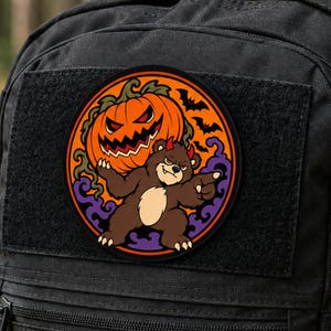 Puede incluir: Un parche circular que muestra un oso pardo con pequeños cuernos rojos señalando hacia adelante, frente a una gran calabaza naranja con una sonrisa dentada. El fondo incluye humo púrpura y murciélagos negros sobre un círculo naranja.