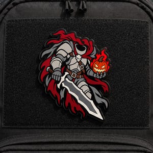 Puede incluir: Una mochila táctica negra con un parche de PVC de un caballero sin cabeza con armadura gris y capa roja, sosteniendo una espada grande en una mano y una calabaza de Halloween naranja brillante en la otra, sobre un fondo de tela negra.
