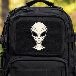 Peut inclure: Un sac à dos tactique noir arborant un écusson central de couleur crème représentant une tête d'extraterrestre aux grands yeux sombres en amande et aux contours faciaux subtils. Le sac comporte plusieurs compartiments zippés et des sangles.