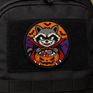 Puede incluir: Un parche circular sobre una superficie de tela negra que muestra un mapache de dibujos animados vestido de vampiro. El mapache sostiene un cubo de dulces con forma de calabaza lleno de golosinas sobre un fondo naranja y morado con murciélagos.