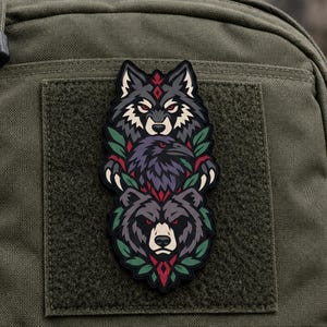 May include: A vertical morale patch featuring a stacked illustration of a wolf, a raven, and a bear head. The design incorporates red, black, and grey tones with green leaf accents. The patch is attached to a dark olive green fabric surface.