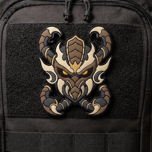 May include: A black, tan, and brown tactical morale patch featuring a stylized scorpion-like creature with glowing yellow eyes. The patch is attached to a black fabric surface with a hook-and-loop fastener, displaying intricate segmented armor details.