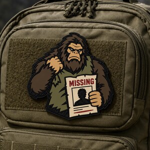 Könnte beinhalten: Ein olivgrüner taktischer Rucksack mit einem rechteckigen Aufnäher, der eine Cartoon-Illustration eines braunen Bigfoot zeigt, der ein weißes Schild mit der Aufschrift MISSING, einer Silhouette und rotem Text hält.