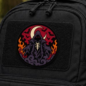 May include: A circular patch attached to a black backpack featuring a hooded figure in a dark robe with glowing red eyes. The background displays a crescent moon, flying bats, orange flames, and purple smoke, all set against a deep red circular backdrop.