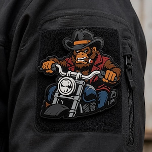 May include: A rectangular morale patch attached to a black sleeve featuring a graphic of a brown gorilla wearing a black cowboy hat and a red jacket while riding a motorcycle. The gorilla has a cigar in its mouth and is gripping the handlebars.