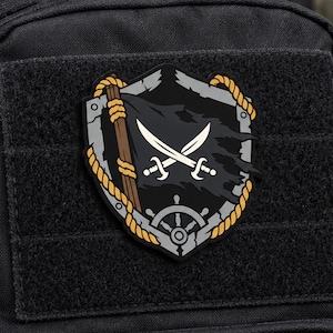 May include: A shield-shaped morale patch featuring a black tattered flag with two crossed white swords, a brown wooden mast, a yellow rope border, and a gray ship's wheel at the bottom, all set against a black hook-and-loop fabric background.