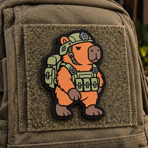 May include: A rubberized morale patch featuring a cartoon capybara wearing a tactical olive green helmet, a matching vest with multiple pouches, and a backpack. The capybara has orange fur and brown paws, set against a dark olive green fabric background.