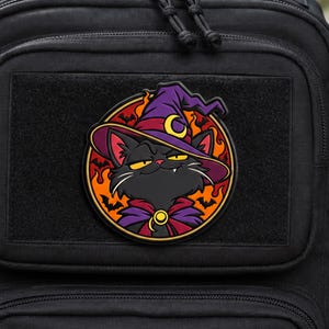 Peut inclure: Un écusson moral circulaire fixé sur un sac à dos tactique noir. L'écusson représente un chat noir portant un chapeau de sorcière violet avec un croissant de lune jaune, une cape violette avec une attache dorée, sur fond de flammes orange et de chauves-souris noires.