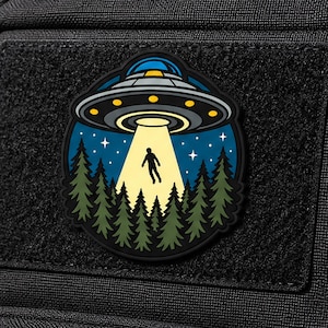 May include: A circular rubber patch featuring a gray UFO with yellow lights hovering over a dark green pine forest. A bright yellow beam of light extends from the craft, illuminating a silhouette of a person floating against a dark blue starry sky.