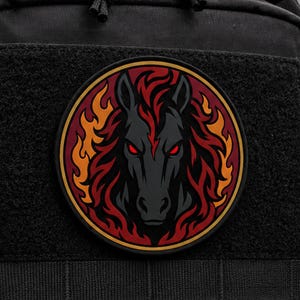 Può includere: Una toppa circolare nera raffigurante una testa di cavallo stilizzata con occhi rossi luminosi e una criniera composta da fiamme arancioni e rosso scuro. Il disegno è racchiuso in un sottile bordo dorato su una superficie in tessuto nero.
